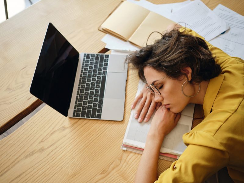 Top view of young tired woman dreamy fall asleep on desk with laptop and documents under head at workplace