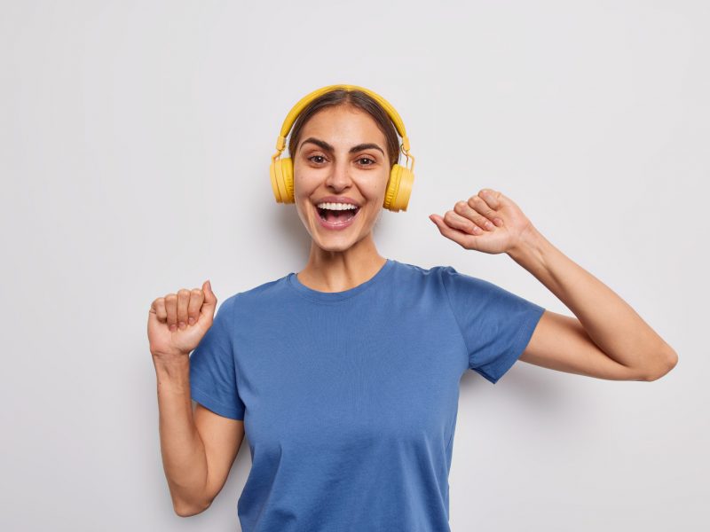 Indoor shot of positive carefree young woman shakes arms moves with rhythm of music laughs joyfully listens music via wireless headphones isolated over white background catches every bit of song